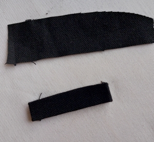 Image shows in irregular rectangle of fabric above a similar piece of fabric that has been folded and pressed into a tab, similar in design to a belt loop