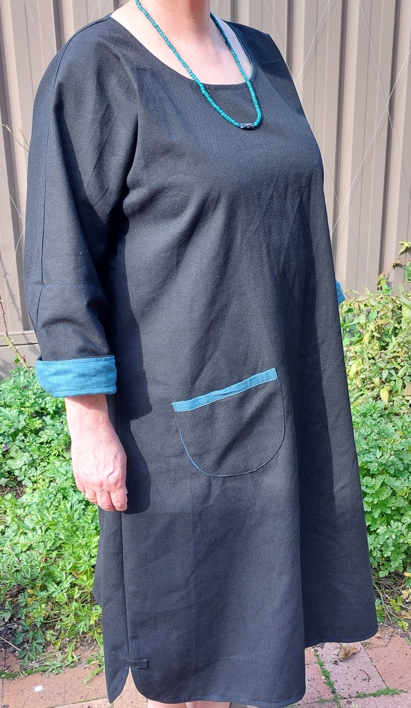 A woman is wearing a black dress with folded up sleeves and a patch pocket. A seam runs from the neck down the arm to the cuff. The hem is curved at the sides and the side seam has a little fabric tab sewn at the bottom to prevent the side seam ripping open under the strain of wear.