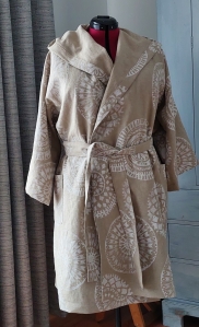 Image shows a belted robe on a dressmakers mannequin. The robe has a shawl collar that extends into a hood. The robe is made out of a coarsely woven cotton fabric in a hessian colour and is printed with large white circular motifs.